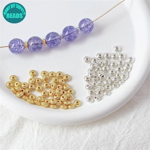 Puede incluir: Primer plano de dos montones de pequeñas cuentas redondas de metal. Un montón es dorado y el otro es plateado. Las cuentas tienen un diseño decorativo en relieve alrededor del borde. Las cuentas están sobre una superficie blanca.