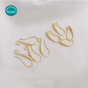 May include: Gold-toned earring hooks, a type of jewellery finding, are arranged on a white surface. The hooks are made of metal and have a smooth, polished finish.