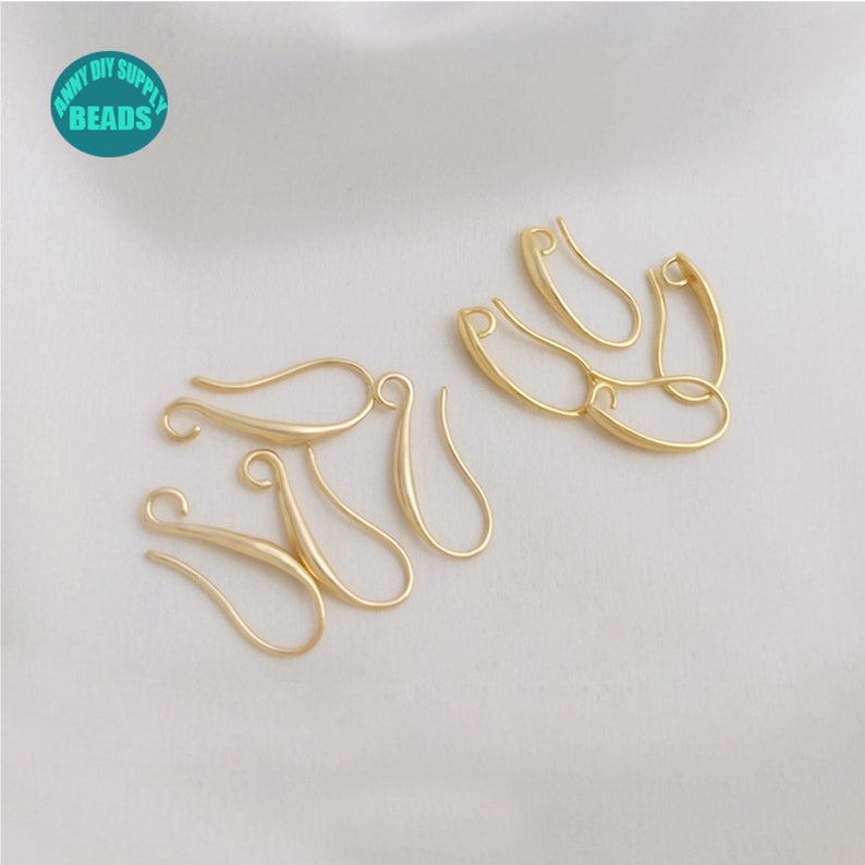 May include: Gold-toned earring hooks, a type of jewellery finding, are arranged on a white surface. The hooks are made of metal and have a smooth, polished finish.