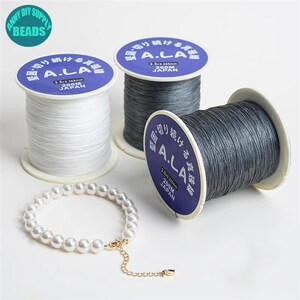 Puede incluir: Tres carretes de hilo gris y blanco etiquetados "A.LA" en japon&eacute;s. Los carretes tienen 200 metros de largo y est&aacute;n hechos en Jap&oacute;n. Un collar de perlas con un cierre de oro est&aacute; en primer plano.