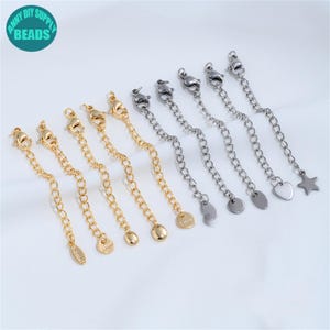 Puede incluir: Una variedad de extensores de cadena de joyería en tonos dorados y plateados. Los extensores presentan cierres de langosta, eslabones de cadena y dijes decorativos, incluyendo una estrella, un corazón y un óvalo. La imagen incluye el texto "ANNY DIY SUPPLY BEADS" en un círculo turquesa.
