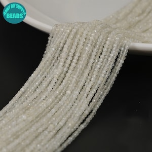 Op de afbeelding: Een streng witte maansteen kralen, elke kraal is gefacetteerd en heeft een licht iriserende glans. De kralen zijn geregen op een doorzichtige draad en zijn ongeveer 2 mm in diameter.