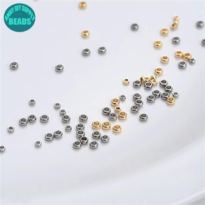 May include: A close-up of a variety of small, round metal jump rings. The jump rings are silver, gold, and black in colour. They are scattered on a white surface.