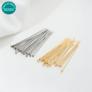 May include: A selection of silver and gold-coloured head pins, suitable for jewellery making and crafts. The pins are straight with a small, flat head. The image includes a teal and white circular logo that reads "ANNY DIY SUPPLY BEADS."