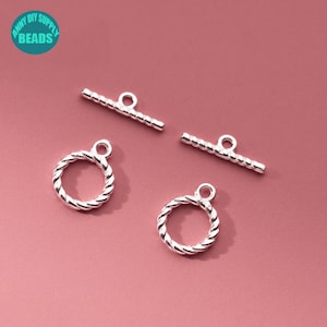 Peut inclure: Des fermoirs de bijoux argentés, comprenant deux anneaux torsadés en forme de corde et deux barres à bascule, sont présentés sur un fond rose. Ces fermoirs sont conçus pour la fabrication de bijoux et comportent de petites boucles.
