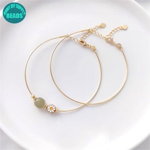Puede incluir: Dos pulseras de brazalete doradas con una delicada extensión de cadena. Una pulsera presenta una cuenta verde y un dije de margarita blanca.