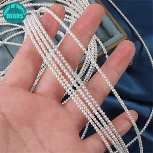 Può includere: Più fili di piccole perle bianche sono tenuti in una mano. Le perle sono infilate insieme, adatte per collane o bracciali. L'immagine include il testo "ARMY DIY SUPPLY BEADS" in un design circolare.