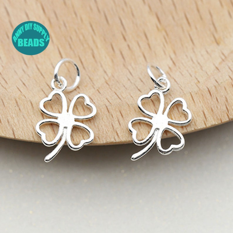 May include: Two silver four-leaf clover charms with a jump ring for attaching to jewelry. The charms are on a light brown wooden surface.