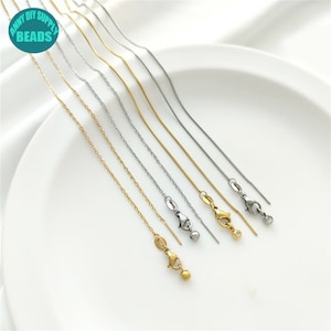 May include: Six different chain styles in gold and silver, each with a lobster clasp and a small ball at the end. The chains are arranged in a row on a white surface.