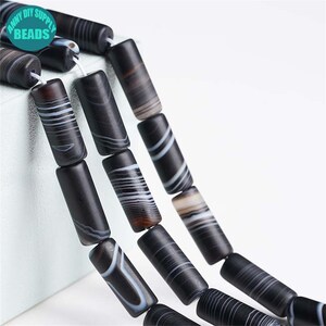 May include: Close-up of black and white striped barrel beads strung together. The beads are cylindrical with white and grey stripes. The image includes a teal circle with the text "ANNY DIY SUPPLY BEADS".
