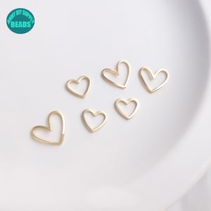 May include: Six small, gold-colored heart-shaped charms are scattered on a white surface. The charms are open-frame designs, with a simple, elegant outline. The upper left corner of the image features a teal circle with the text "RAINY DIY SUPPLY BEADS".