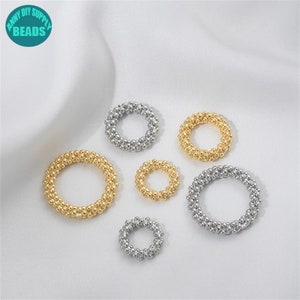 Puede incluir: Una variedad de anillos de cuentas doradas y plateadas de varios tamaños. Los anillos están dispuestos sobre una superficie blanca y sedosa. Los anillos están hechos de pequeñas cuentas redondas, creando una apariencia texturizada. La imagen incluye un logotipo que dice "ANNY DIY SUPPLY BEADS".