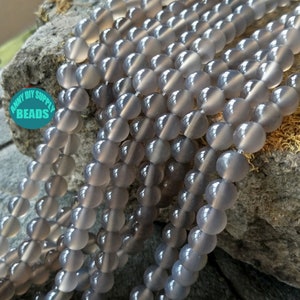 May include: A strand of grey agate beads. The beads are round and smooth, with a slightly translucent appearance. The beads are arranged in a loose pile on a grey rock.