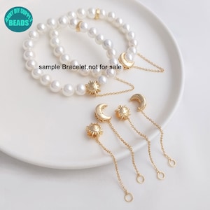 May include: A white pearl bracelet with gold sun and moon charms. The bracelet is on a white surface with three additional gold sun and moon charms on chains.