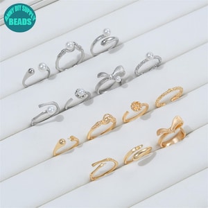 Könnte beinhalten: Eine Sammlung von silber- und goldfarbenen Ringen, die in Reihen angeordnet sind. Die silbernen Ringe haben Perlenakzente, Schleifenformen und offene Bänder. Die goldenen Ringe umfassen strukturierte Bänder, Schleifendesigns und offene Fassungen.