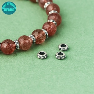 May include: Three silver bead caps with a decorative design. The bead caps are on a green surface. A bracelet with red and clear beads is in the background.