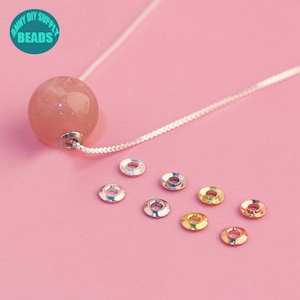May include: A close-up of a variety of jump rings in silver, gold, and rose gold. The jump rings are arranged in a circle on a pink surface. A pink stone bead is hanging from a silver chain in the background.