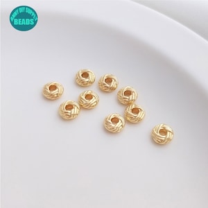 Puede incluir: Cuentas redondas doradas con textura tejida dispersas sobre una superficie blanca. Cada cuenta tiene un pequeño agujero central, ideal para la creación de joyas o manualidades. La imagen incluye un logotipo que dice "ANNY DIY SUPPLY BEADS".