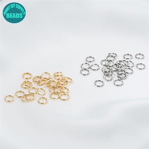 May include: A selection of small, gold and silver jump rings, scattered on a white surface. The jump rings are circular and appear to be metal. The image also includes a teal circle with the text "ANNY DIY SUPPLY BEADS".
