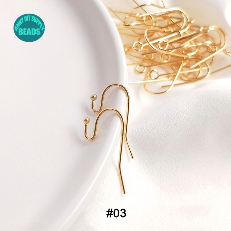 May include: Two gold-toned earring hooks with a small ball at the top of each hook. The hooks are on a white surface. #03