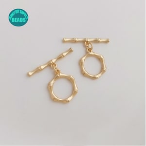 Pode incluir: Dois fechos de correr de bambu em tom dourado com um anel redondo preso a cada um. Os fechos são de metal e têm um design de bambu.
