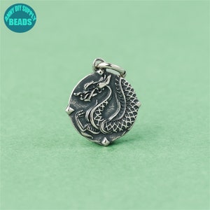 May include: Silver charm featuring a detailed dragon design. The charm is round and has a loop at the top for attaching to a necklace or bracelet.