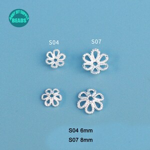 May include: Four silver-colored, flower-shaped beads. Two are labeled S04, measuring 6mm, and two are labeled S07, measuring 8mm. The beads have a delicate, openwork design. The background is a solid light blue.