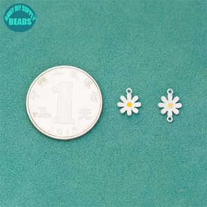 K&ouml;nnte beinhalten: Zwei charmartige Anh&auml;nger in Form einer G&auml;nseblume mit wei&szlig;en Bl&uuml;tenbl&auml;ttern und gelben Zentren, neben einer M&uuml;nze. Die Anh&auml;nger haben oben eine kleine Schlaufe zum Befestigen an Schmuck. Der Hintergrund ist t&uuml;rkisgr&uuml;n. Der Text "ANNY DIY SUPPLY BEADS" befindet sich in einem Kreis oben links.