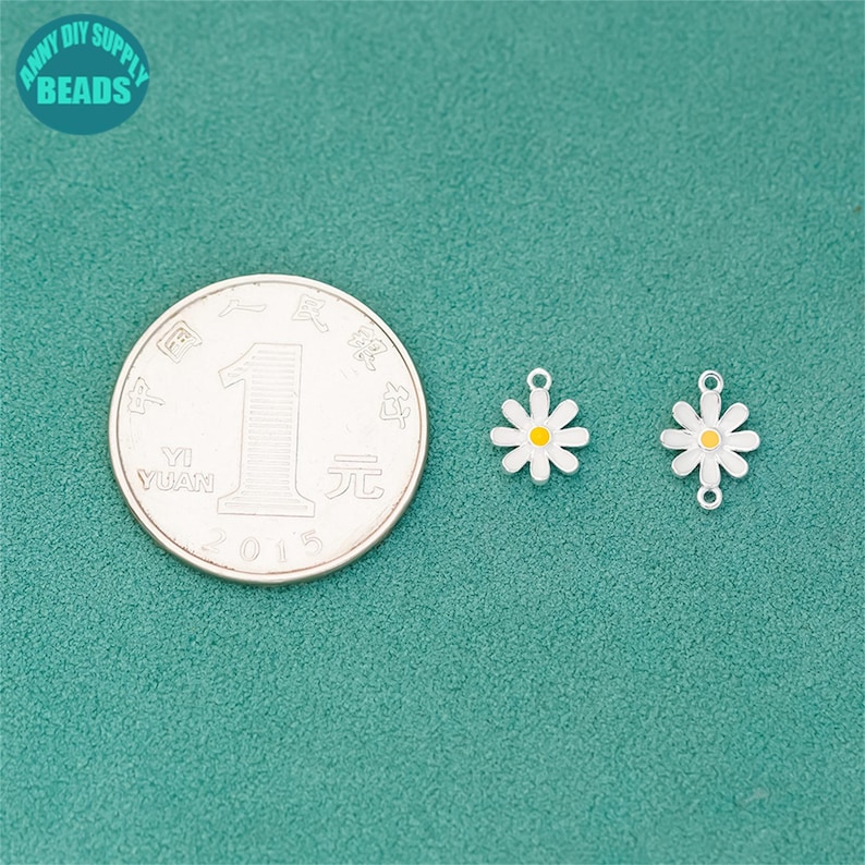 K&ouml;nnte beinhalten: Zwei charmartige Anh&auml;nger in Form einer G&auml;nseblume mit wei&szlig;en Bl&uuml;tenbl&auml;ttern und gelben Zentren, neben einer M&uuml;nze. Die Anh&auml;nger haben oben eine kleine Schlaufe zum Befestigen an Schmuck. Der Hintergrund ist t&uuml;rkisgr&uuml;n. Der Text "ANNY DIY SUPPLY BEADS" befindet sich in einem Kreis oben links.