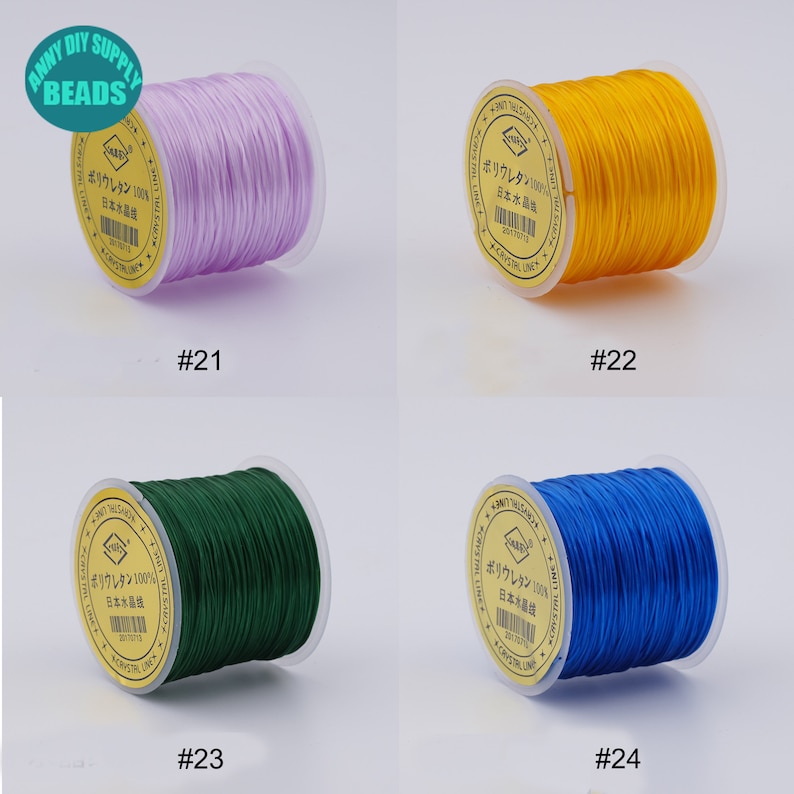 May include: Four spools of elastic cord in different colours: purple, yellow, green, and blue. Each spool has a label with Japanese text and the number 100% on it. The spools are labelled with numbers 21, 22, 23, and 24.