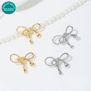 Puede incluir: Cuatro dijes en forma de lazo, dos dorados y dos plateados, con un collar de perlas en el fondo. Los dijes tienen un diseño en bucle con extremos en forma de lágrima. La esquina superior izquierda tiene el texto "ANNY DIY SUPPLY BEADS".