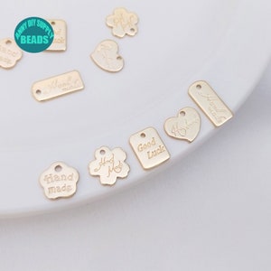 Può includere: Assortimento di ciondoli in metallo dorato con vari disegni e iscrizioni, tra cui "Hand Made", "Good Luck" e "Handmade".