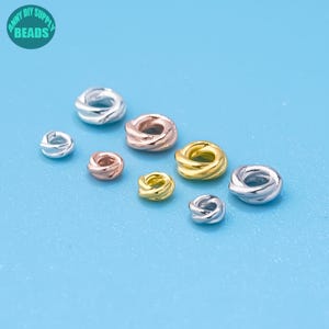 May include: An assortment of twisted metal beads in silver, rose gold, and gold tones. The beads vary in size and are scattered on a light blue surface. The image includes a circular logo with the text "ANNY DIY SUPPLY BEADS".