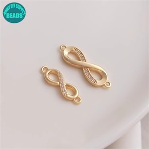 Könnte beinhalten: Zwei goldene Unendlichkeits-Anhänger mit klaren Kristall-Akzenten. Die Anhänger haben an jeder Seite eine Schlaufe zum Anbringen an Schmuck.