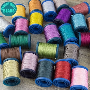 Könnte beinhalten: Sortiment an bunten Metallic-Garnrollen mit blauen Kunststoffspulen. Die Rollen sind auf einer Holzoberfläche angeordnet. Der Text "Anny DIY Supply Beads" ist in der oberen linken Ecke des Bildes sichtbar.