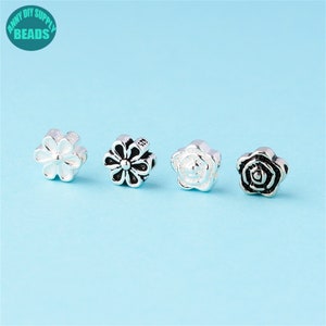 May include: Four silver flower charms, two are daisies and two are roses. The daisies are silver with black centres, and the roses are silver with black centres.