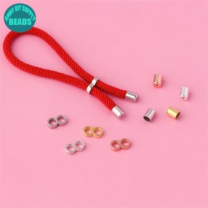 Puede incluir: Un cordón rojo con tapas de extremo plateadas y conectores de bucle infinito plateados, dorados y de oro rosa. El cordón está sobre una superficie rosa.
