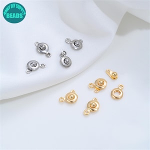 May include: A collection of metal jewelry findings including silver-toned and gold-toned circular snap fasteners. Each piece features a central raised dome and a small loop for attachment, designed for use in jewelry making and craft projects.