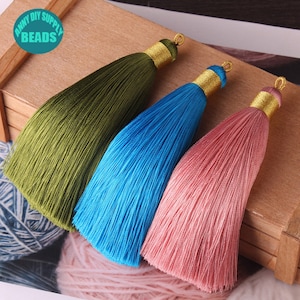 Könnte beinhalten: Drei Seidenquasten in Grün, Blau und Rosa mit goldenen Verzierungen. Die Quasten liegen auf einer Holzoberfläche.
