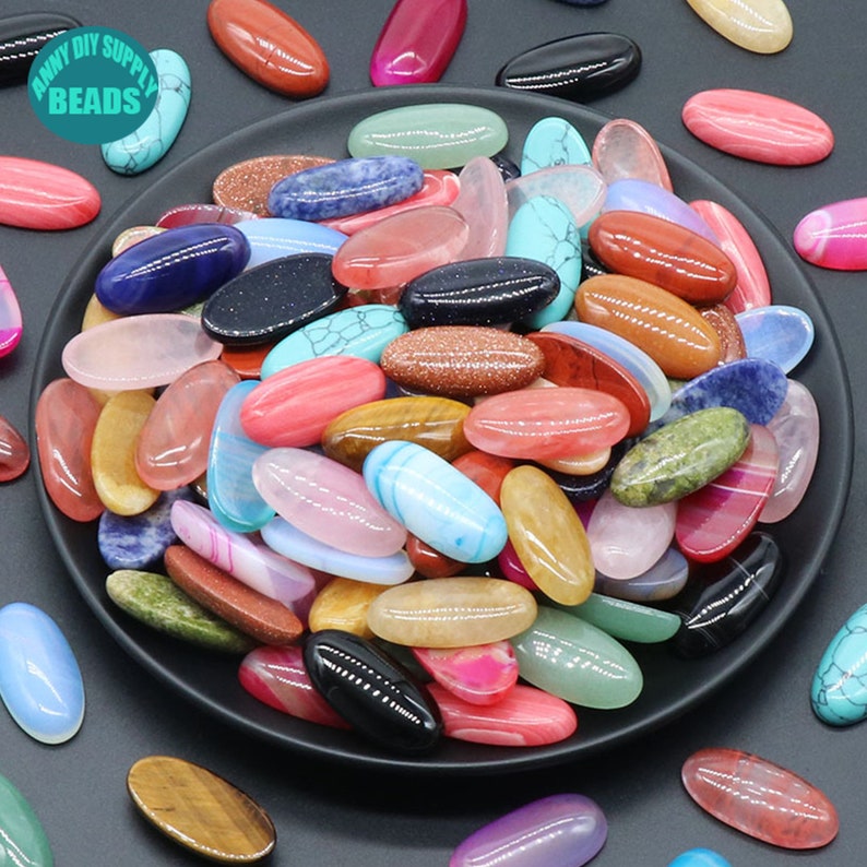 Pu&ograve; includere: Una collezione di cabochon ovali in pietra preziosa, lucidati in vari colori, tra cui rosso, rosa, blu, verde, marrone e bianco. Le pietre sono disposte in una ciotola su una superficie nera.