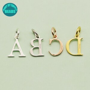 May include: Four silver and gold letter charms, A, B, C, and D, with a silver loop at the top. The charms are on a light green background. The letters are in a serif font. The charms are stamped with "S925" on the back.