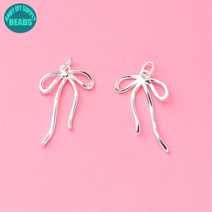 Pode incluir: Dois pingentes de laço de prata com um laço para prender em joias. Os laços são feitos de metal fino e têm um design delicado e elegante.