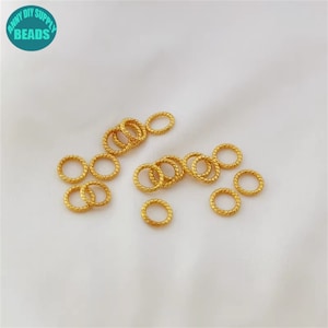 Puede incluir: Un grupo de 20 anillos de salto de metal de color dorado. Los anillos tienen un diámetro de aproximadamente 1,25 cm y presentan un diseño trenzado.