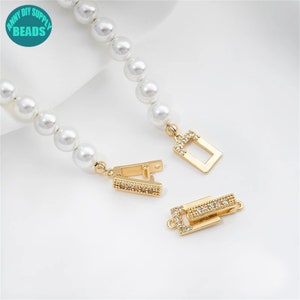 May include: Gold-tone clasp with clear crystal accents for jewelry making. The clasp is made of two pieces, one with a rectangular shape and the other with a hinged design. The clasp is shown with a strand of white pearls.