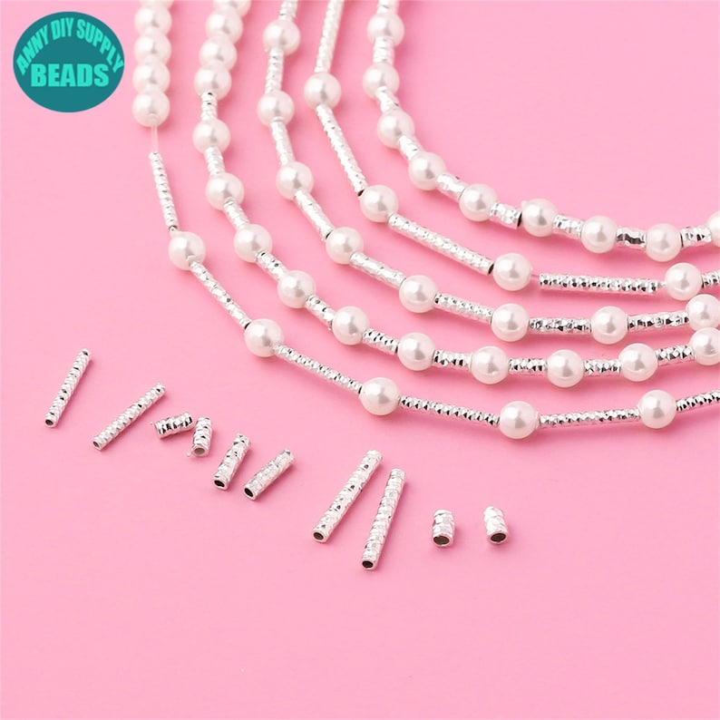 May include: A set of white pearl beads and silver spacer beads. The beads are arranged in a line on a pink surface. There are also some silver spacer beads scattered around the pearls.