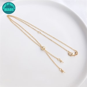 Puede incluir: Un collar dorado con una cadena delicada y una cuenta deslizante. El collar presenta un pequeño cierre y dos colgantes colgantes. La imagen incluye el texto "ANNY DIY SUPPLY BEADS" en un círculo turquesa.