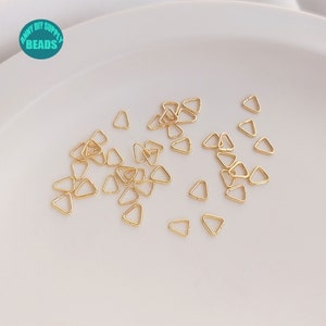 May include: A close-up image of a pile of small, gold-coloured triangle jump rings. The jump rings are scattered on a white surface.