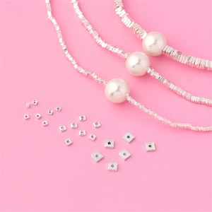 Może przedstawiać: Trzy srebrne łańcuszki z kwadratowymi koralikami i trzy białe perły. Rozrzucone na różowym tle są srebrne kwadratowe kółka do łączenia z otworem w środku.