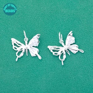 Könnte beinhalten: Zwei silberne Schmetterlingsanhänger mit filigranen Details. Die Schmetterlinge sind in entgegengesetzte Richtungen ausgerichtet und haben ein zartes, durchbrochenes Design.
