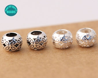 May include: Four silver metal beads with a floral design. The beads are round and have a hole in the centre. The beads are arranged in a row on a brown surface.